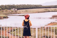 一位戴着无檐帽的女子倚着栏杆，凝望着四周被自然环抱的平静湖泊