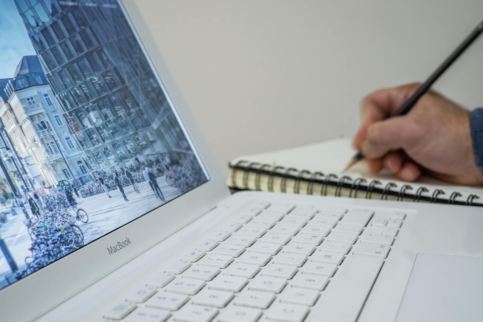 A person taking notes in a notebook beside an open laptop displaying an urban scene.