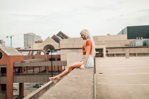 Free Side View Photo of Woman Sitting on Edge of Brown Concrete Building Stock Photo