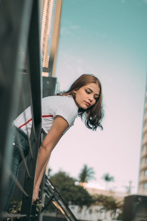 Photo of Woman in White Shirt and Blue Jeans Leaning Forwards on Metal Railing