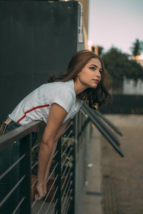 Photo of Woman in White Shirt and Blue Jeans Leaning Forwards on Metal Railing While Looking up