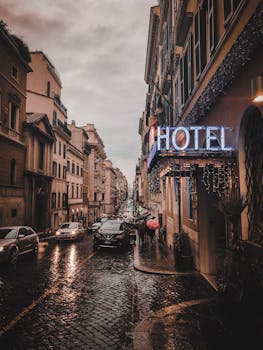 A rainy city street scene at dusk, featuring a glowing neon hotel sign, wet cobblestones, and bustling cars.