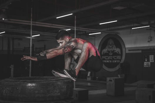 Tattooed man jumping onto large tire indoors at a gym.