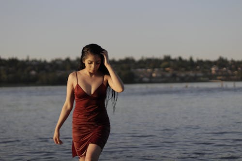Photo of Woman in Red Spaghetti-strap Dress Walking out from Body of Water Looking Down