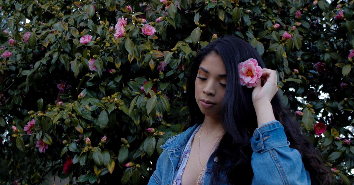 A young woman poses in a garden holding a pink flower against a lush backdrop.