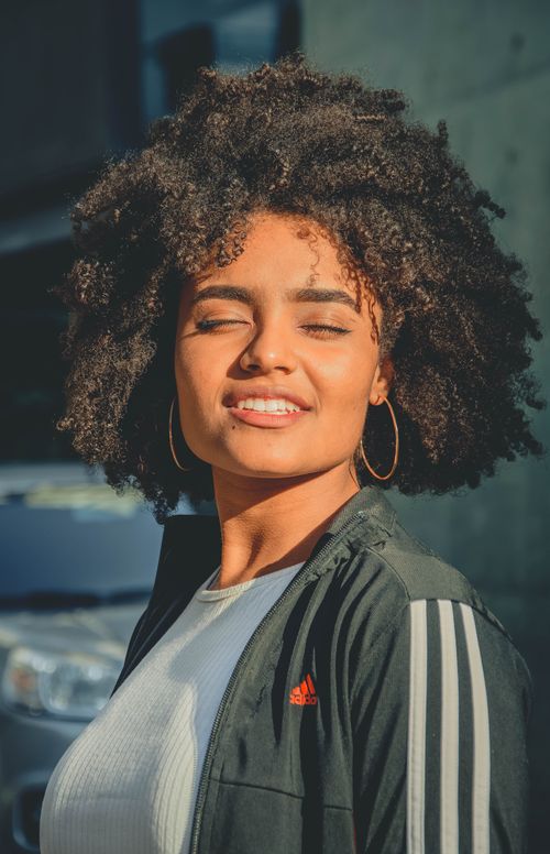 Free Stylish young woman with afro hairstyle smiling in sunlight outdoors. Stock Photo