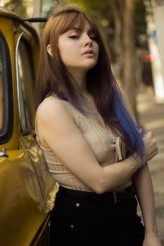 一名留着棕色长发的年轻女子在一辆黄色汽车旁摆出姿势，背景是城市街道