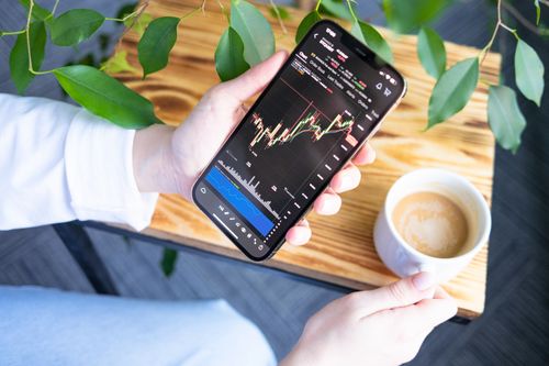 Free Woman holding smartphone showing stock market graph while enjoying a cup of coffee. Stock Photo