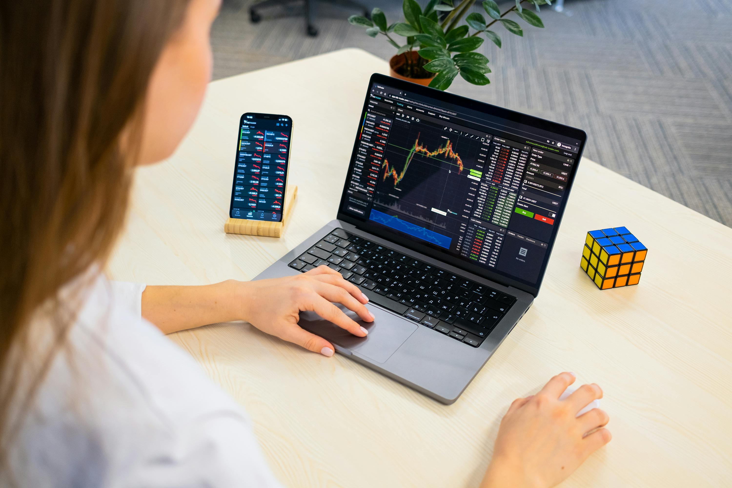 A woman using a laptop for stock trading analysis with a smartphone and Rubik's Cube nearby.