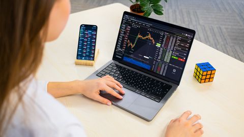 A woman using a laptop for stock trading analysis with a smartphone and Rubik's Cube nearby.