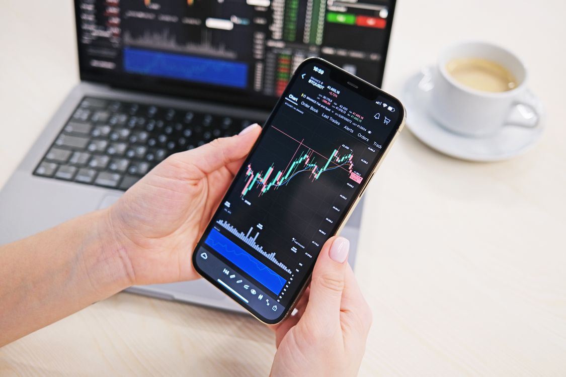 Free Close-up of hands holding smartphone displaying stock charts with laptop in the background on a desk. Stock Photo