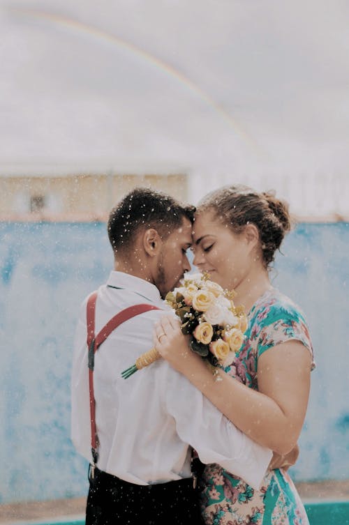 Photo of Man in White Shirt and Suspenders Holding Woman in Floral Dress Holding Flowers while Closing Eyes