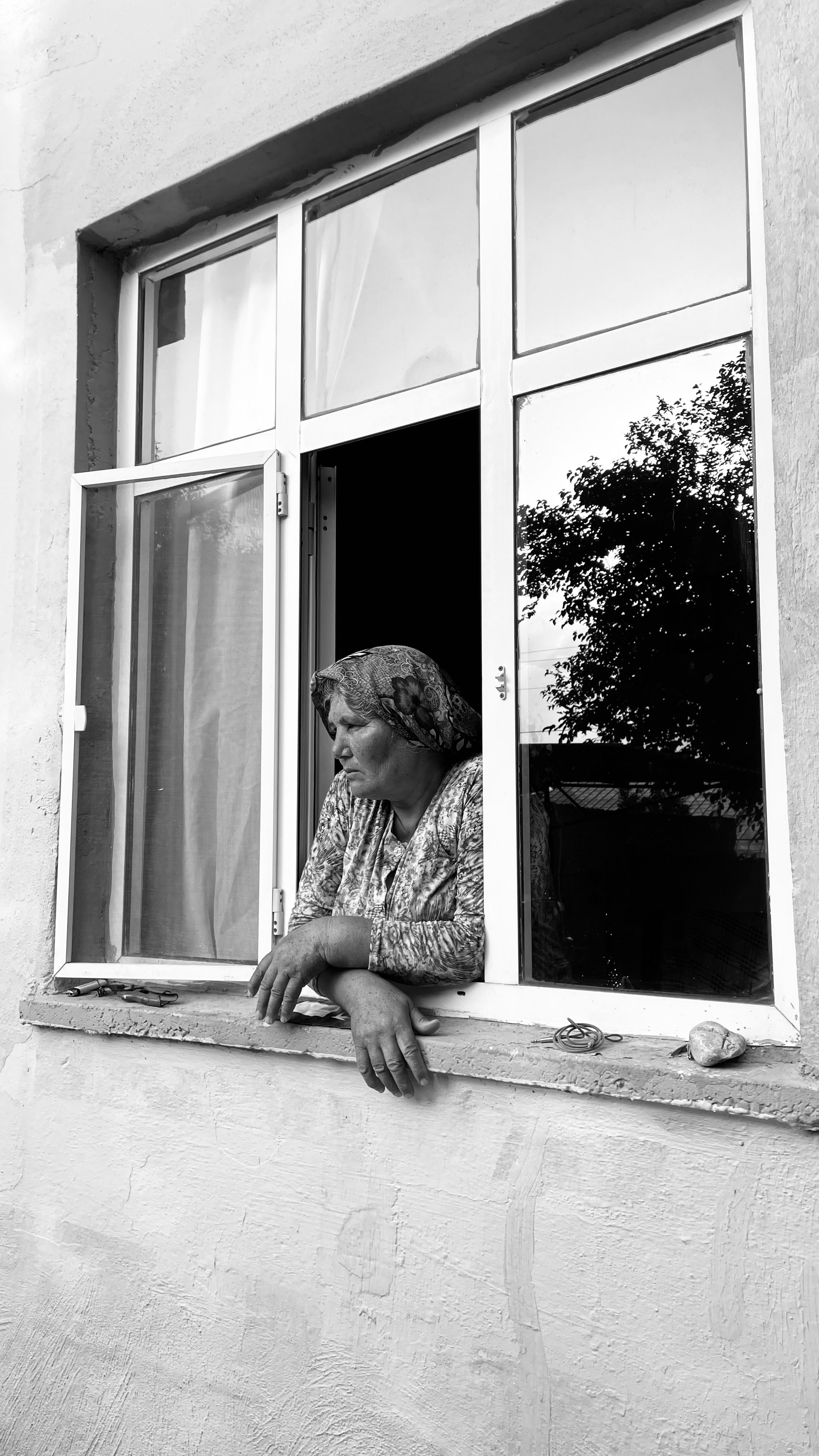 Senior woman with headscarf looking out of window with reflection of trees.