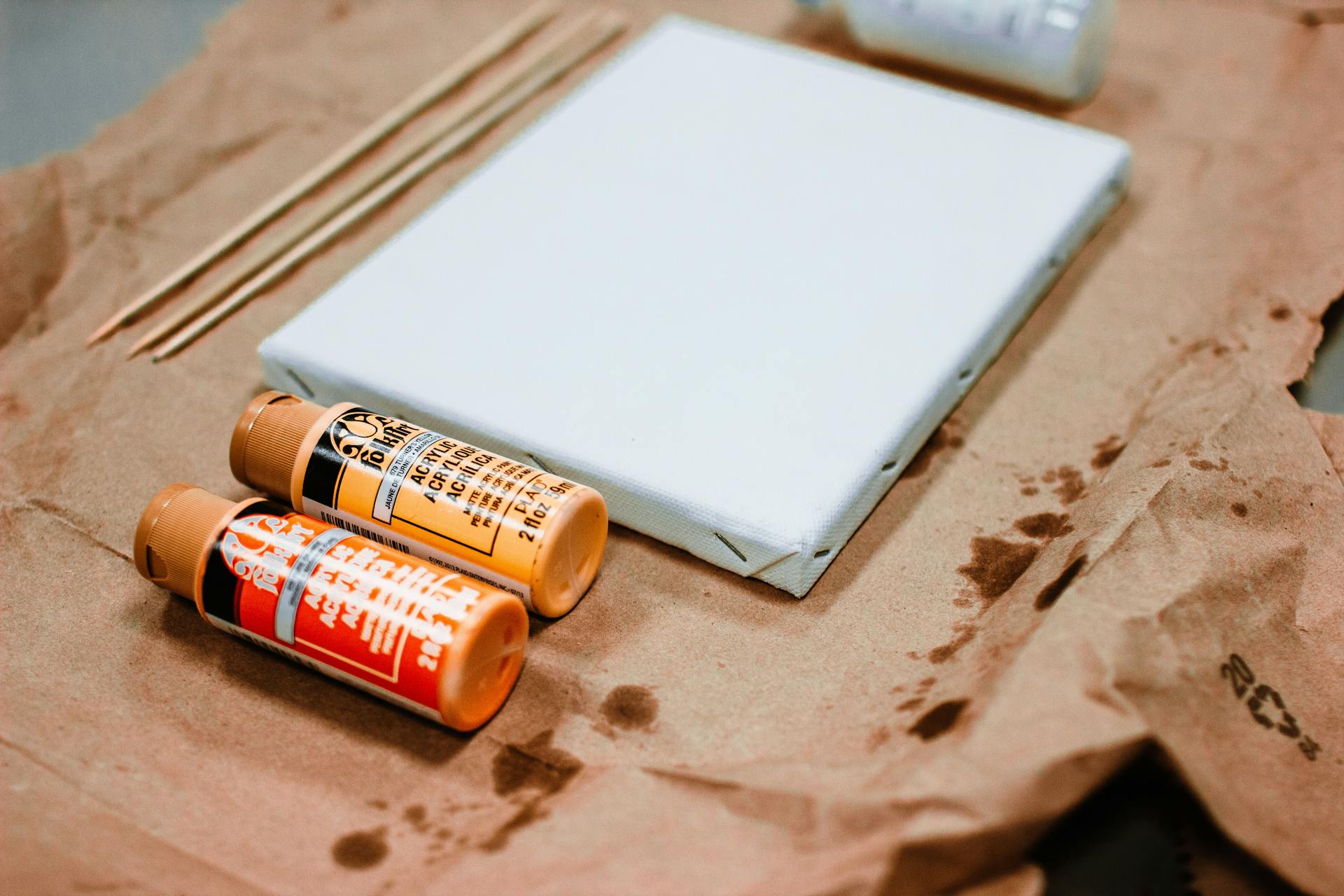 Close-up of art supplies including canvas, paint bottles, and wooden sticks on a craft paper surface.