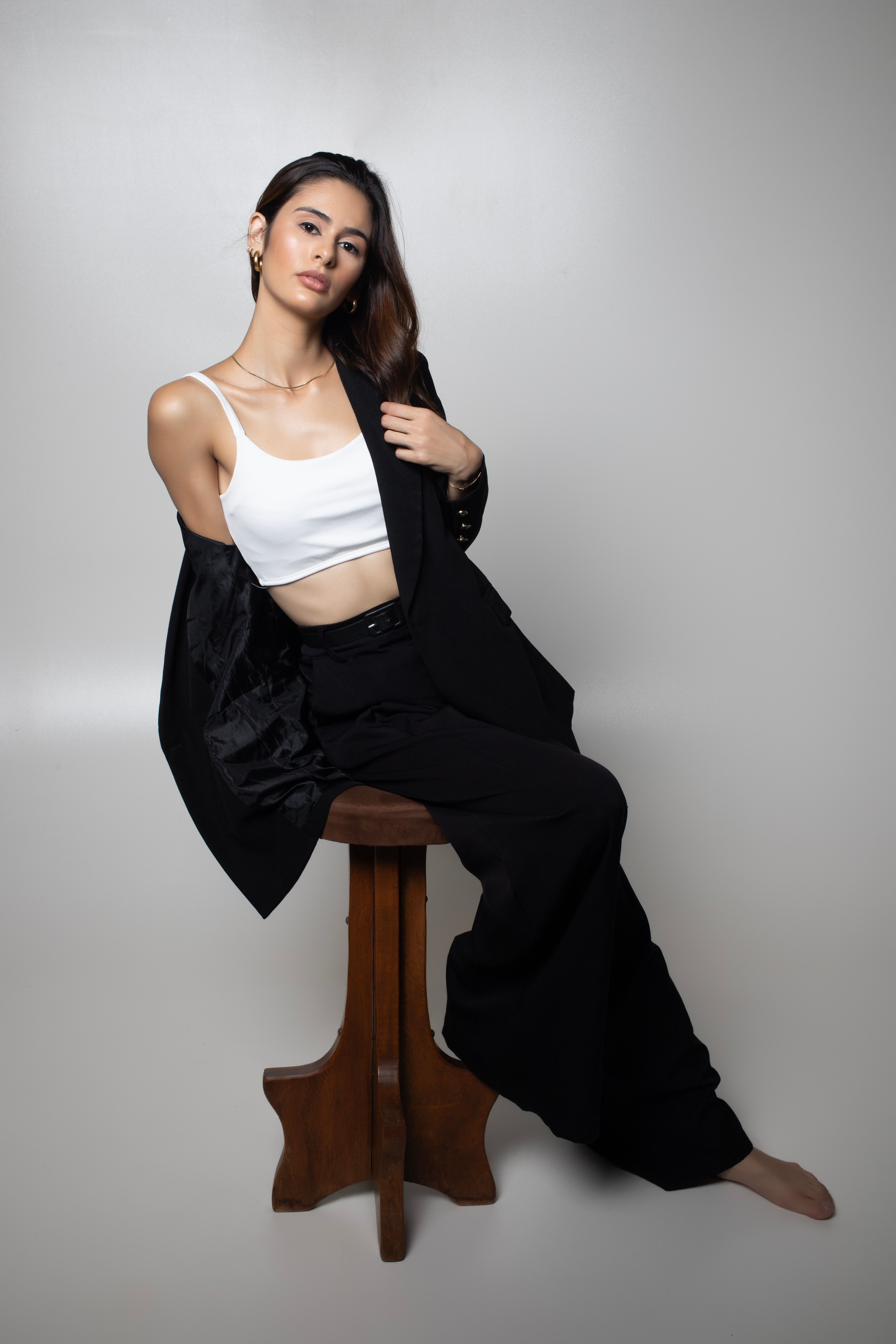 A stylish woman posing in a black suit and white top on a wooden stool, showcasing elegance.