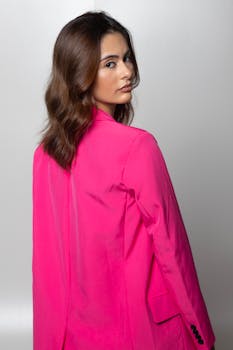 Stylish brunette woman posing confidently in a bright pink jacket against a white studio background.