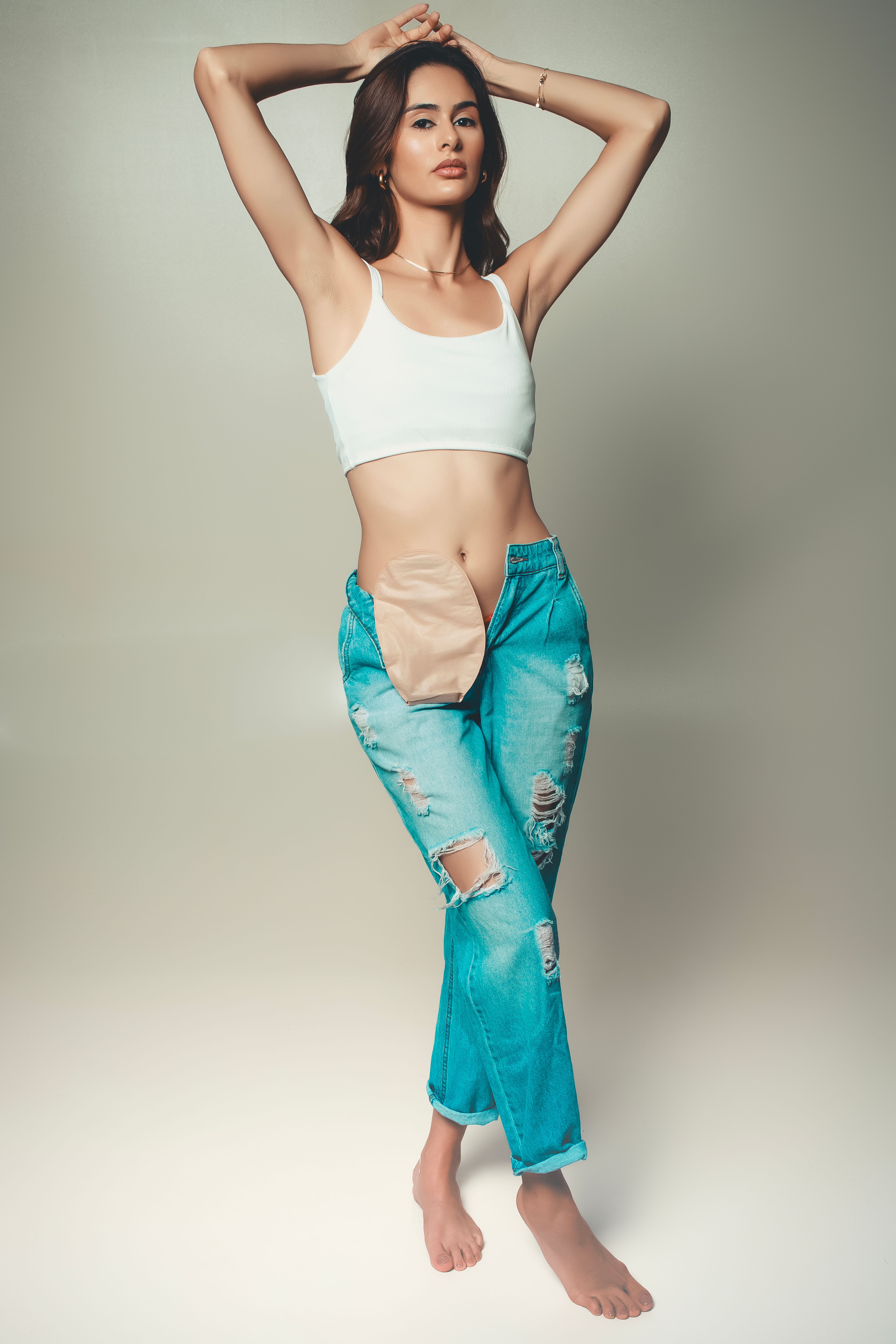 Fashionable woman posing in ripped jeans and cropped top against a simple backdrop.