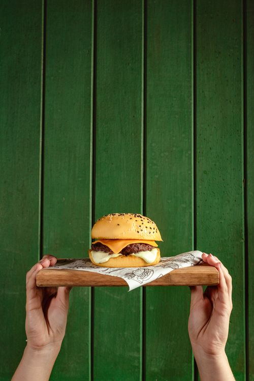 Free Delicious cheeseburger served on a wooden tray held by hands against a vibrant green background. Stock Photo