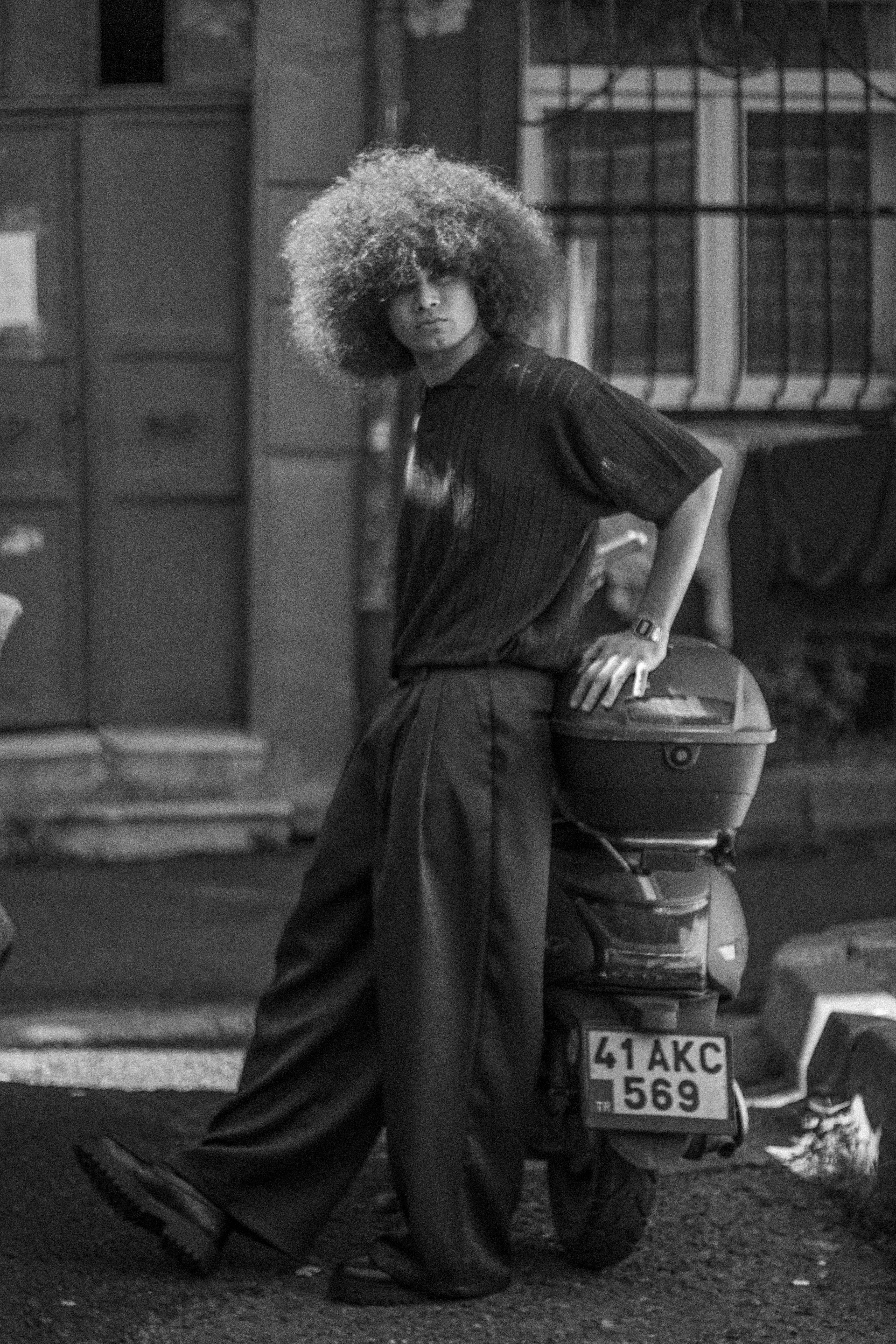 Black and white portrait of a young man with afro hair leaning on a motorcycle in a city street.