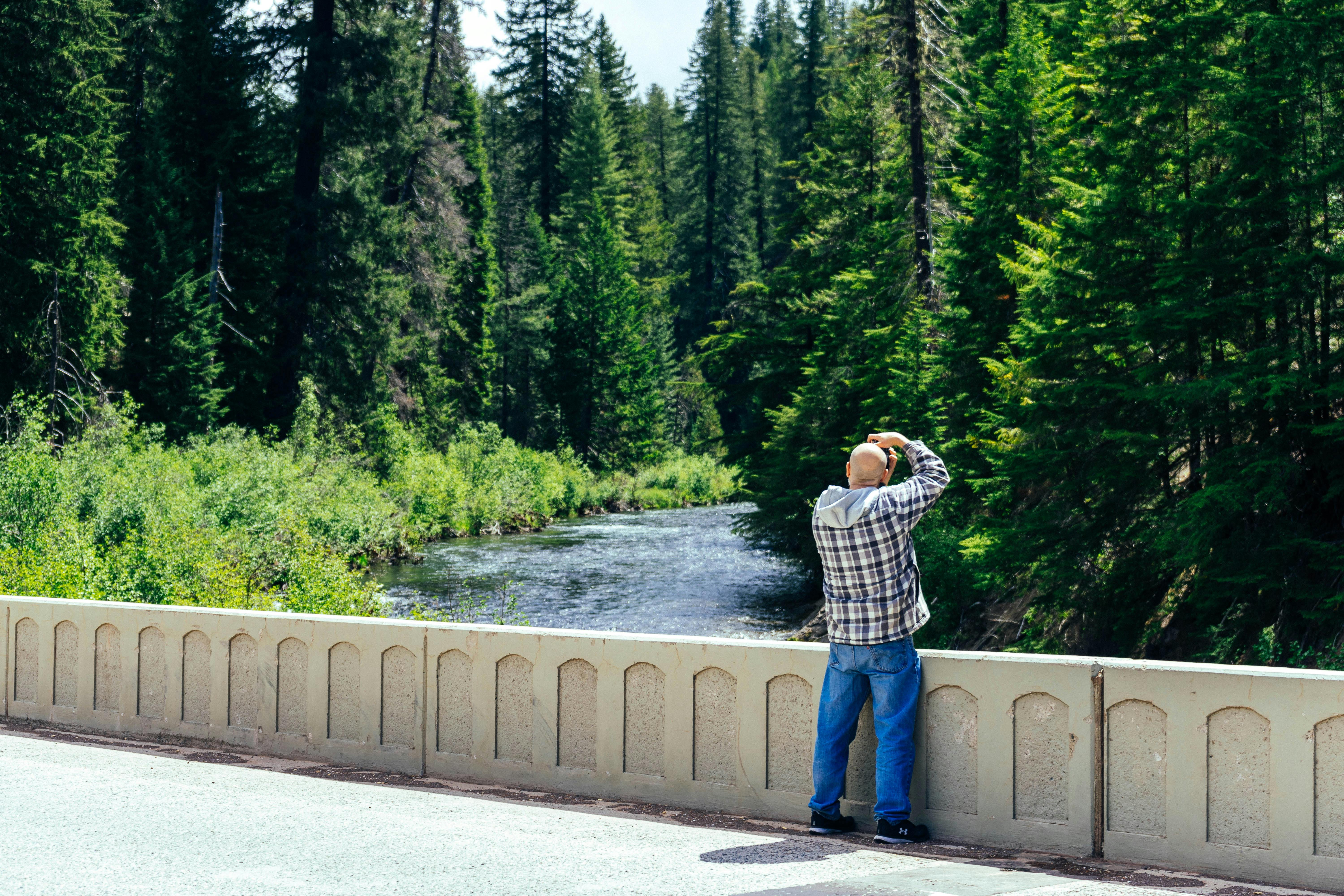 photo of man standing while taking picture of trees and river