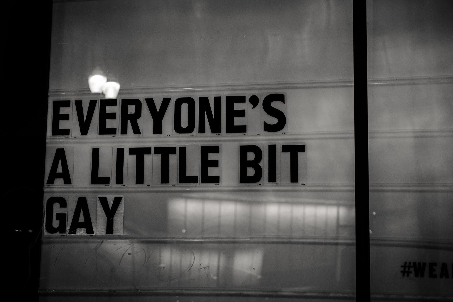 Black and white photo of a sign reading 'Everyone's a little bit gay', promoting inclusivity.
