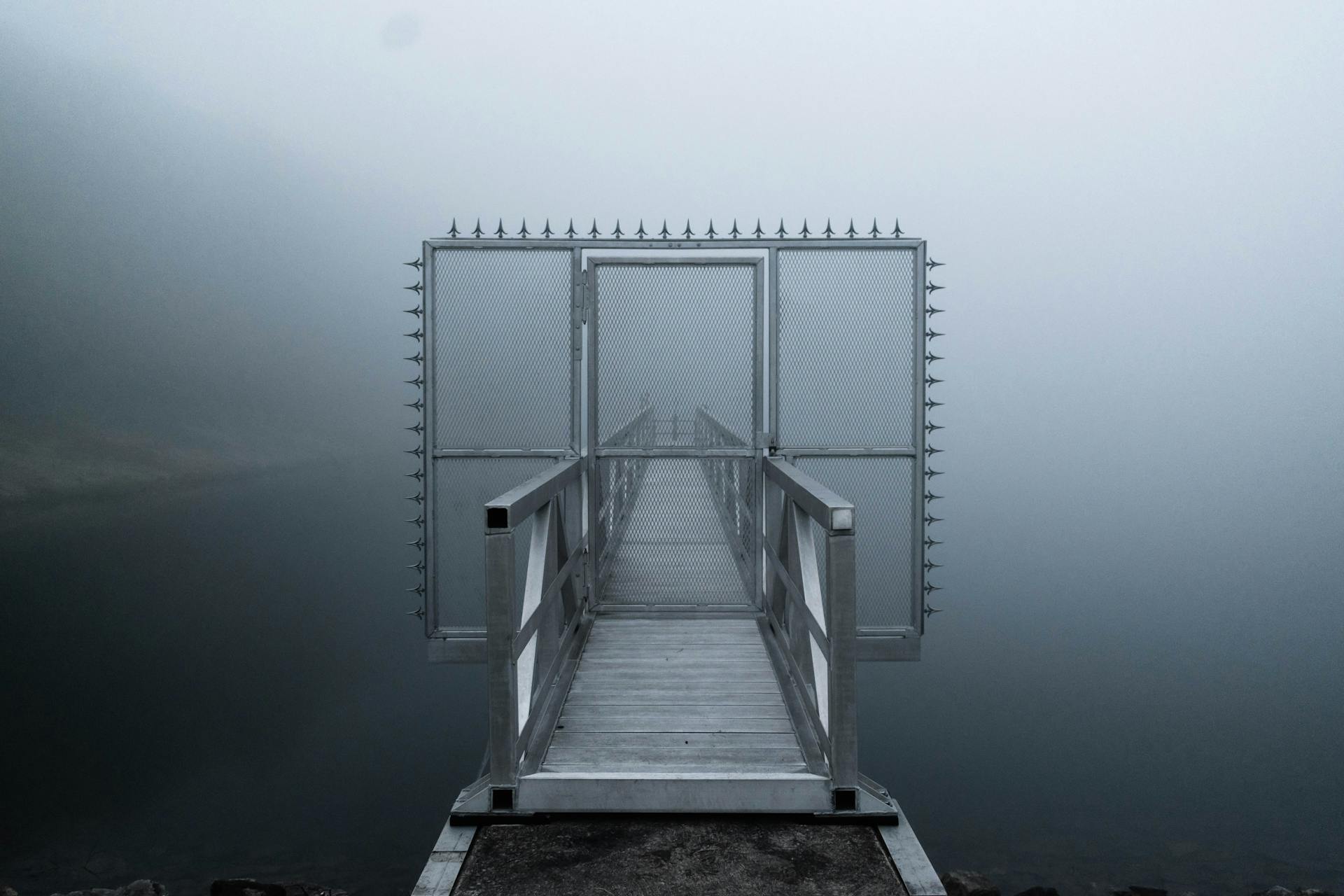 A foggy morning scene with a metal dock leading to a barred gate over tranquil water.
