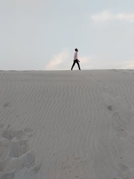 Silhouette of a person walking atop a vast sandy dune under a soft morning sky.