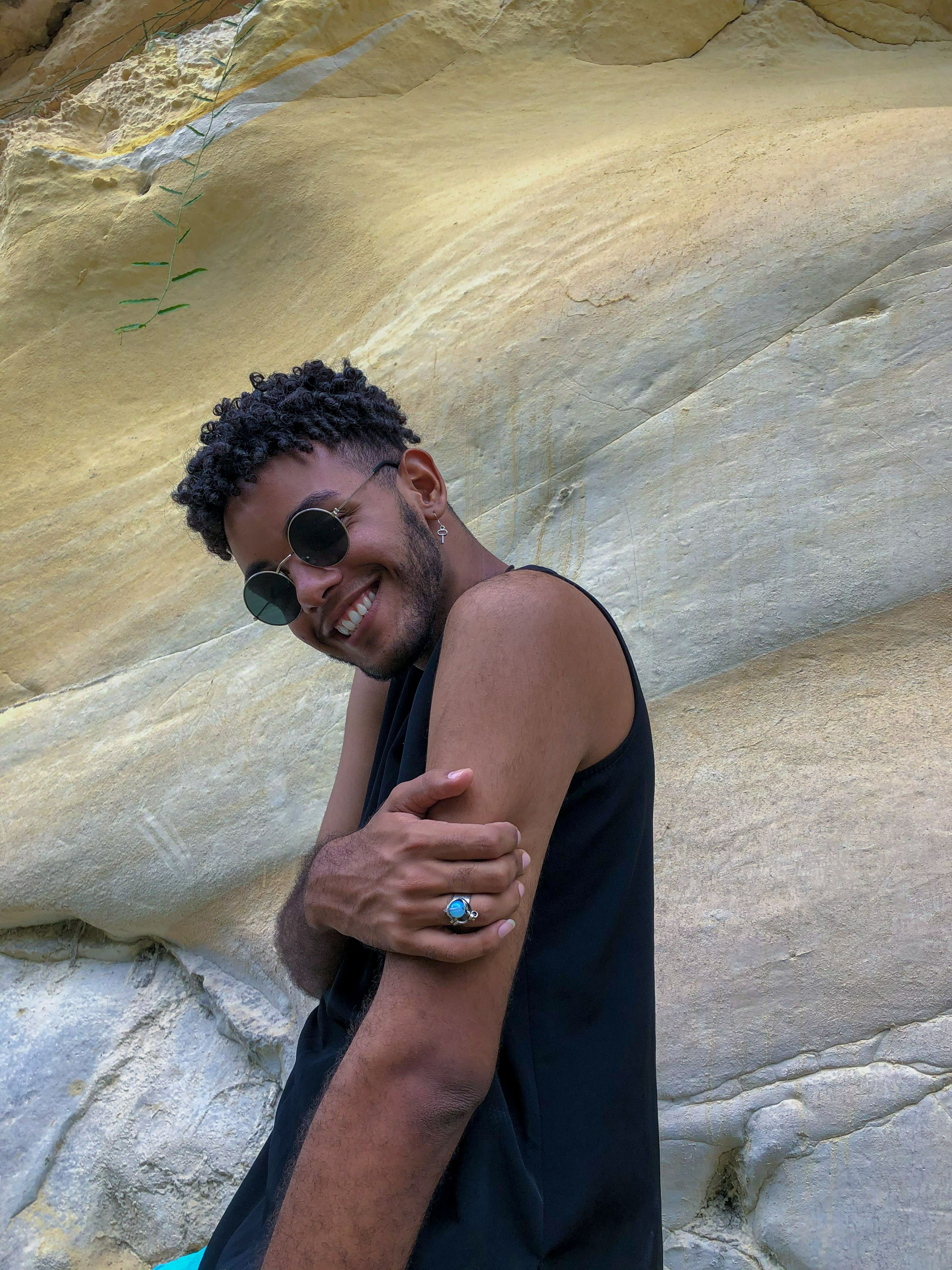 A stylish young man smiling and posing against a rock formation in La Habana, Cuba, showcasing fashion and confidence.