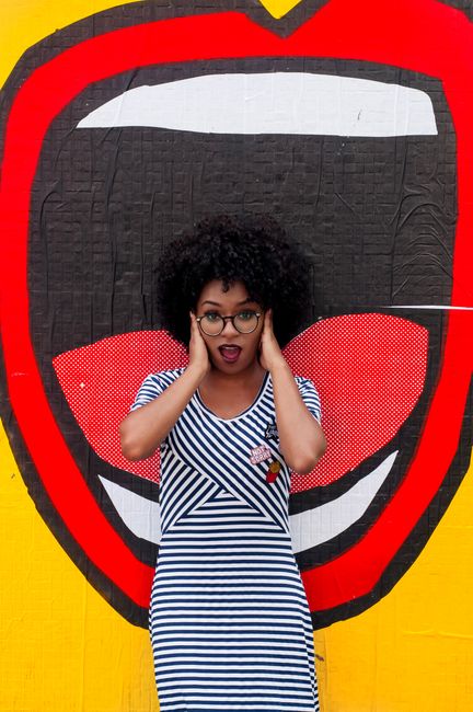 Stylish woman with afro hair poses against a colorful, artistic mural in a striped dress.