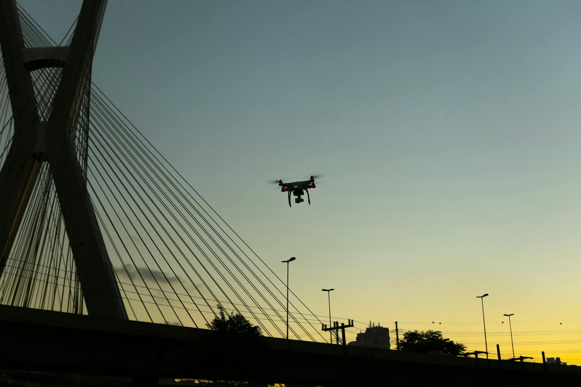 Silhouette of a drone flying at sunset near a modern cable-stayed bridge.