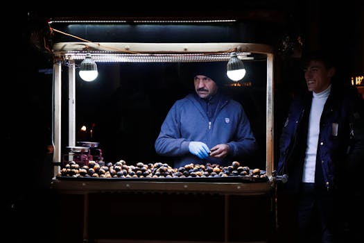 A vendor selling roasted chestnuts on a cart in the street at night, interacting with customers.