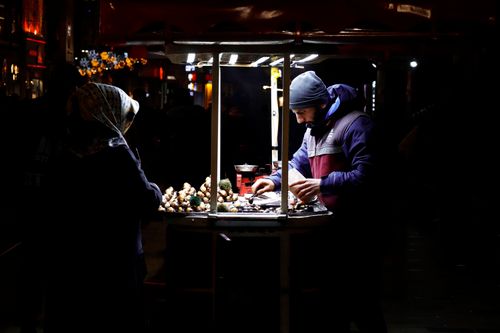 Free A street vendor selling food at night from a lit cart in an urban setting, interacting with a customer. Stock Photo
