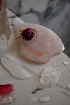 Rose quartz gua sha on marble with flower petals and water droplets, perfect for skincare and relaxation.