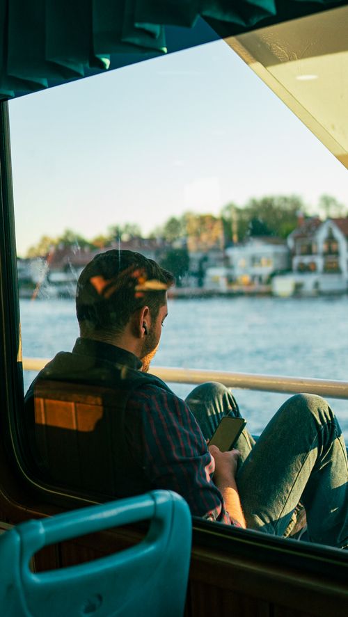 Free Young man sits on a boat, enjoying a peaceful river view under the evening sun. Stock Photo