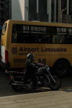 Motorcyclist passes airport bus on bustling South Korean city street during the day.