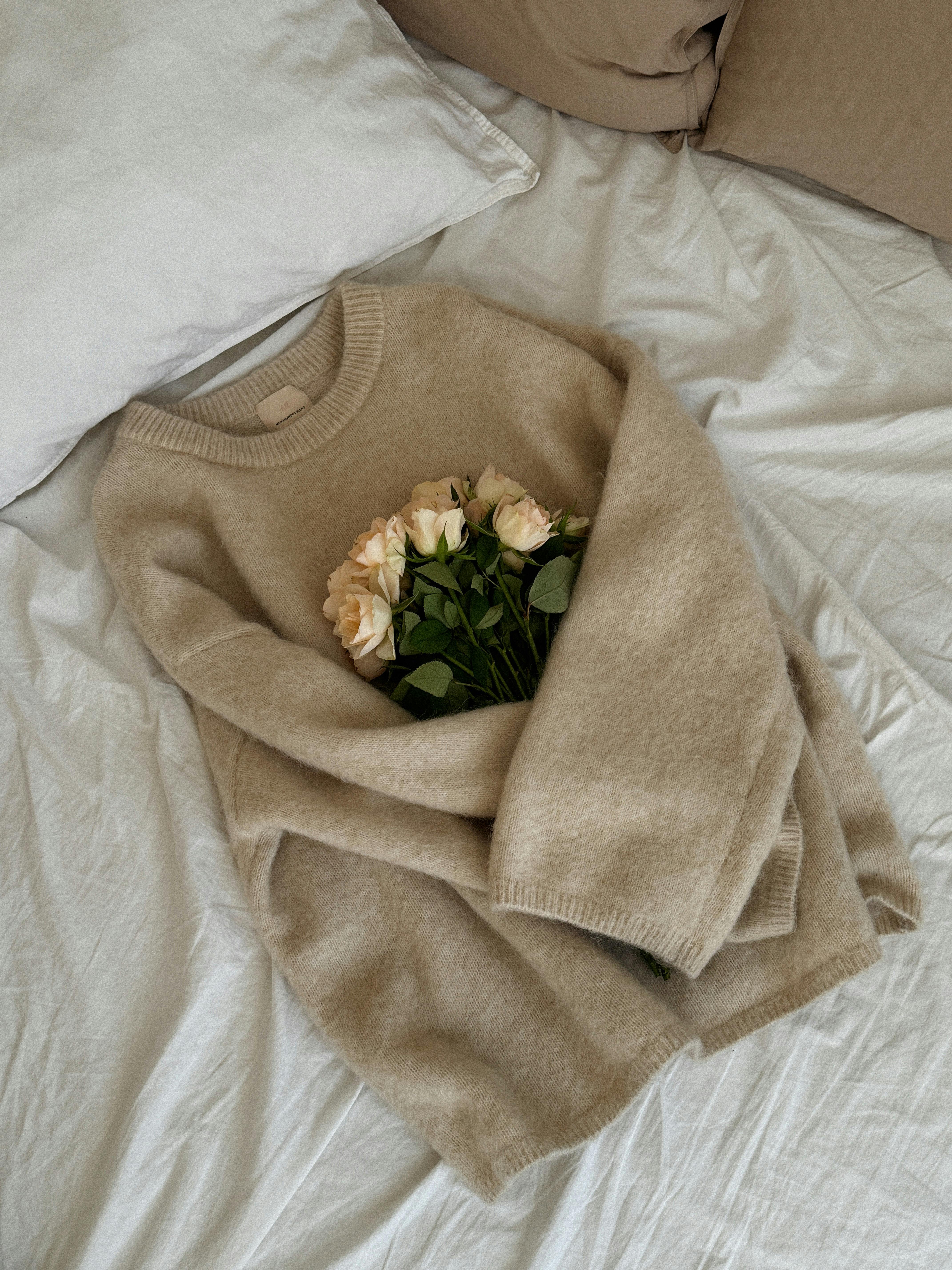 A soft beige sweater holding white roses, placed on a bed for a cozy vibe.