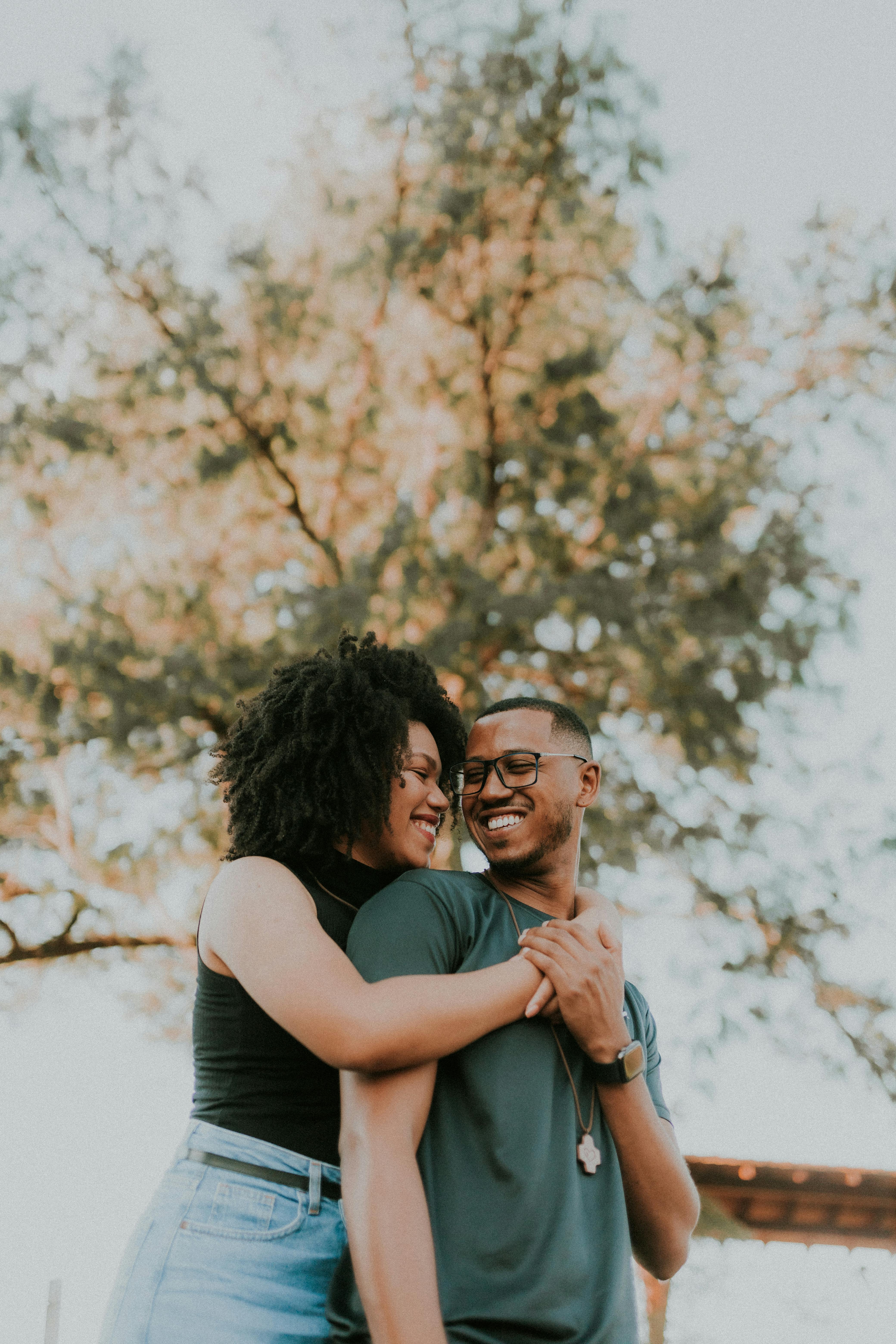 Smiling couple embraces outdoors under a sunlit tree, radiating joy.
