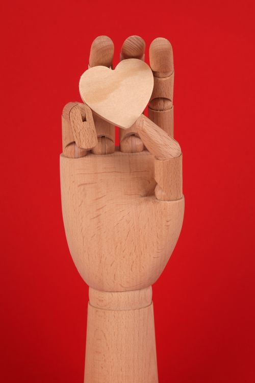 Free A wooden mannequin hand grasping a heart shape against a vibrant red background. Stock Photo