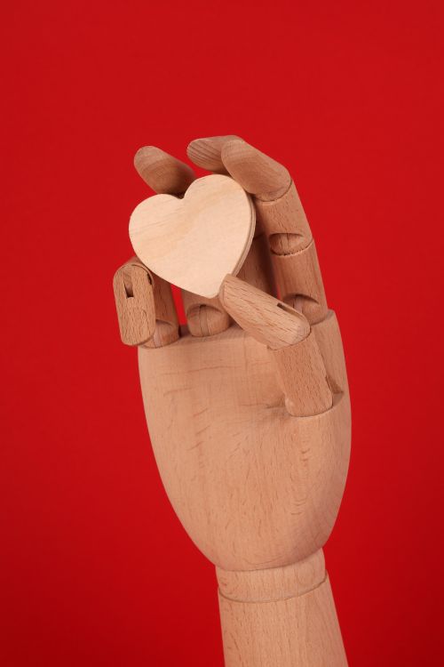 Free A wooden mannequin hand holding a small wooden heart against a vibrant red background. Stock Photo