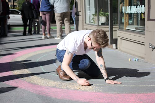 Photo of a Man in White T-Shirt Coloring on Gray Pavement Next to a Building 