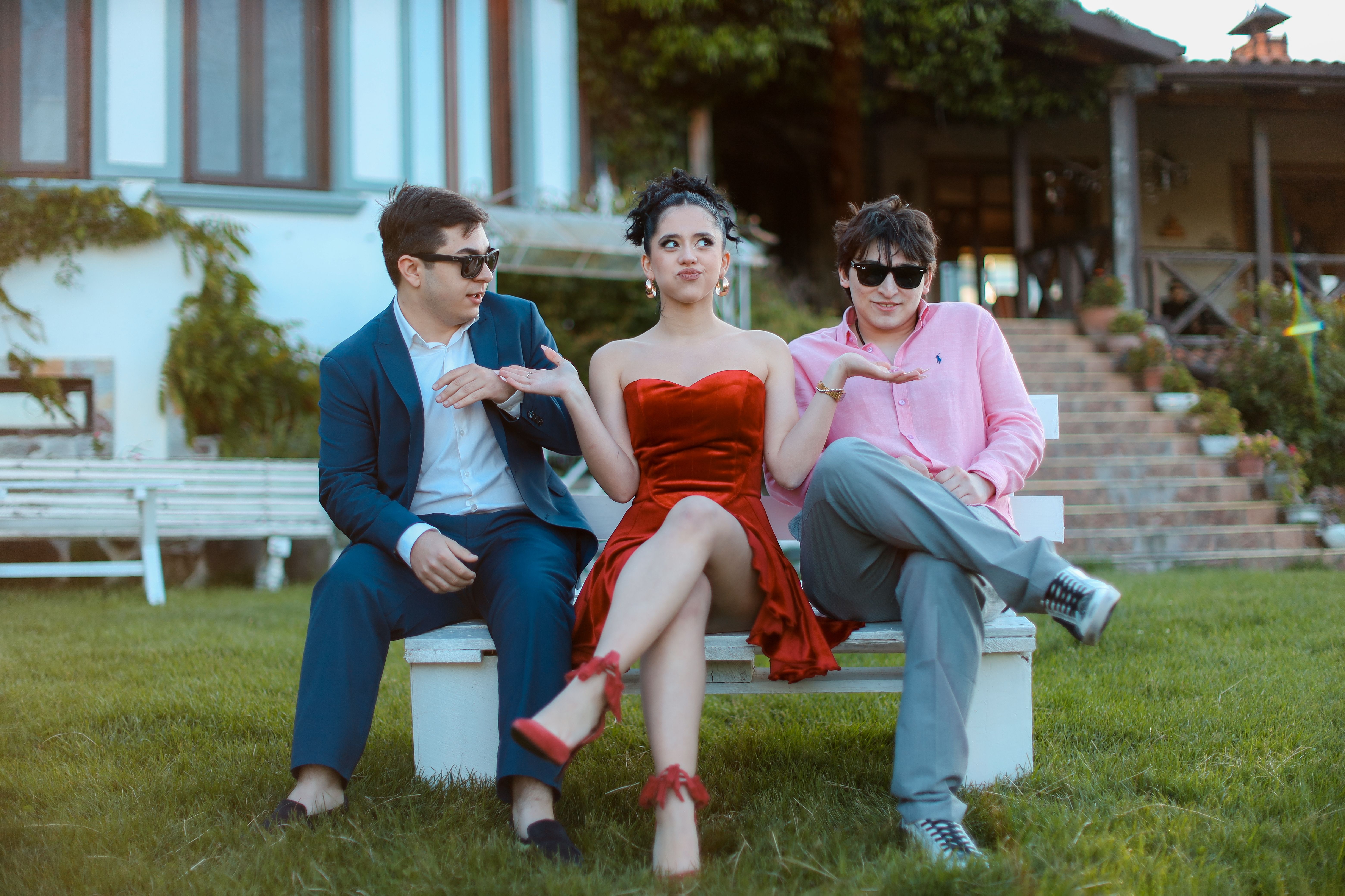 Three young adults in stylish attire sitting together on a bench outdoors, showing camaraderie and elegance.