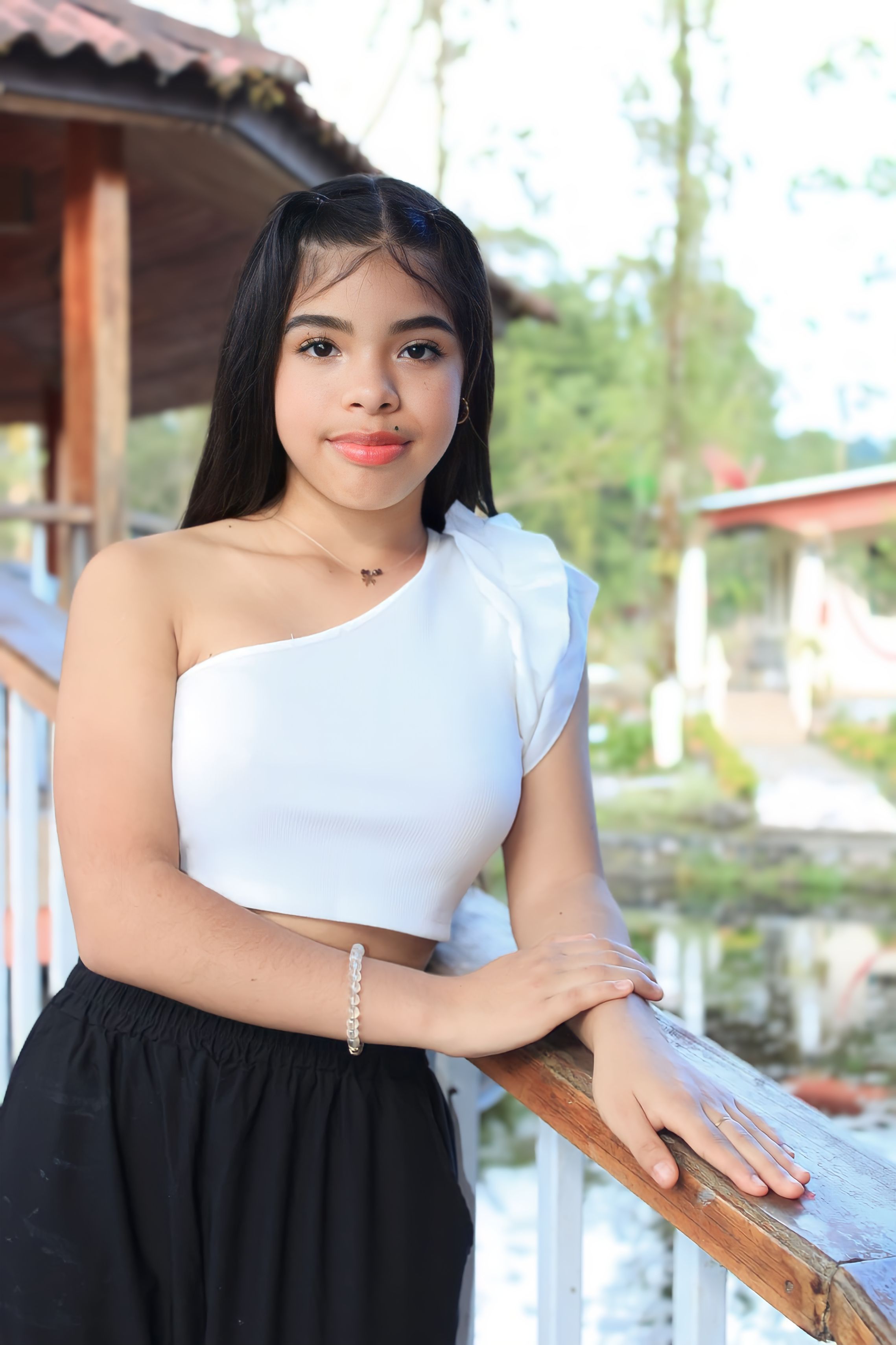 Charming young woman in white top and black pants, posing by railing in an outdoor setting.