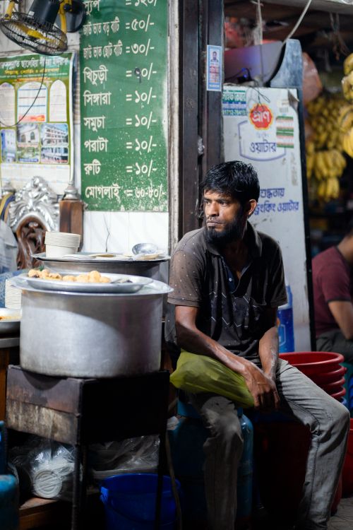 Free A street vendor sits amidst vibrant market life, surrounded by local produce and signs. Stock Photo