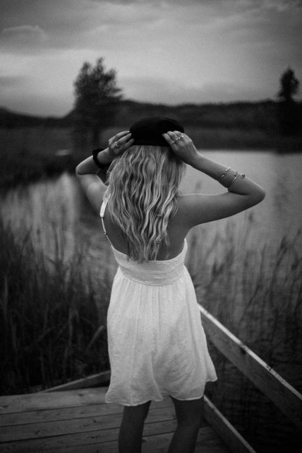 Monochrome image of a woman in a white dress with a hat, standing by a serene lakeside.