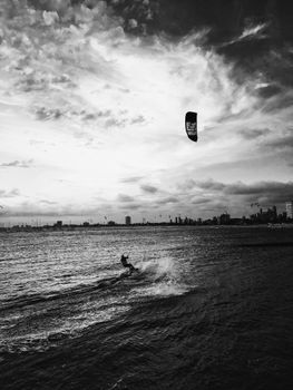 Black and white photo of a kiteboarder riding waves with Melbourne skyline in the background.