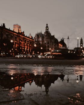 City street view at night with buildings and lights reflecting in puddles after rainfall.