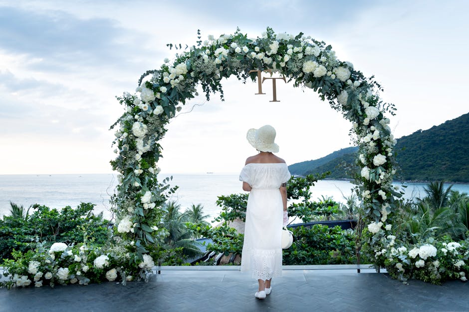 A woman in a white dress stands under a floral arch, overlooking the sea at a wedding.
