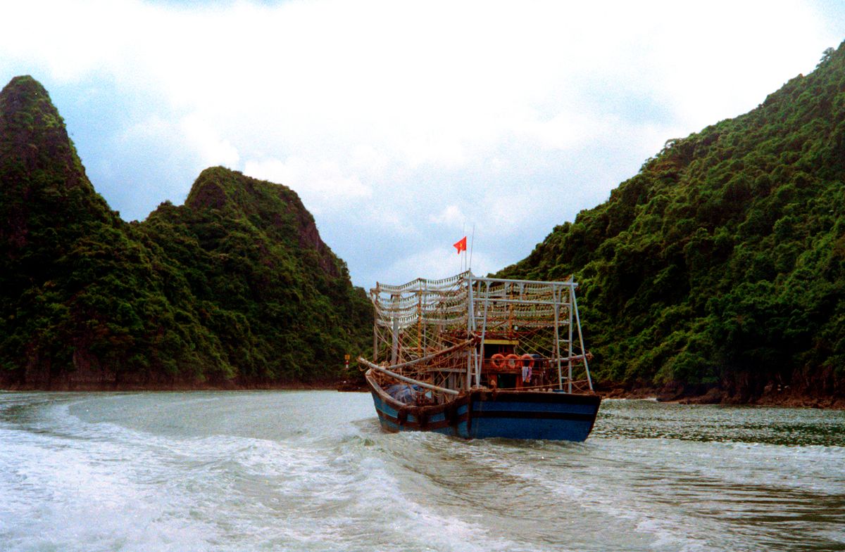 A wooden boat moving between Ha Long Bay limestone islands under a cloudy sky