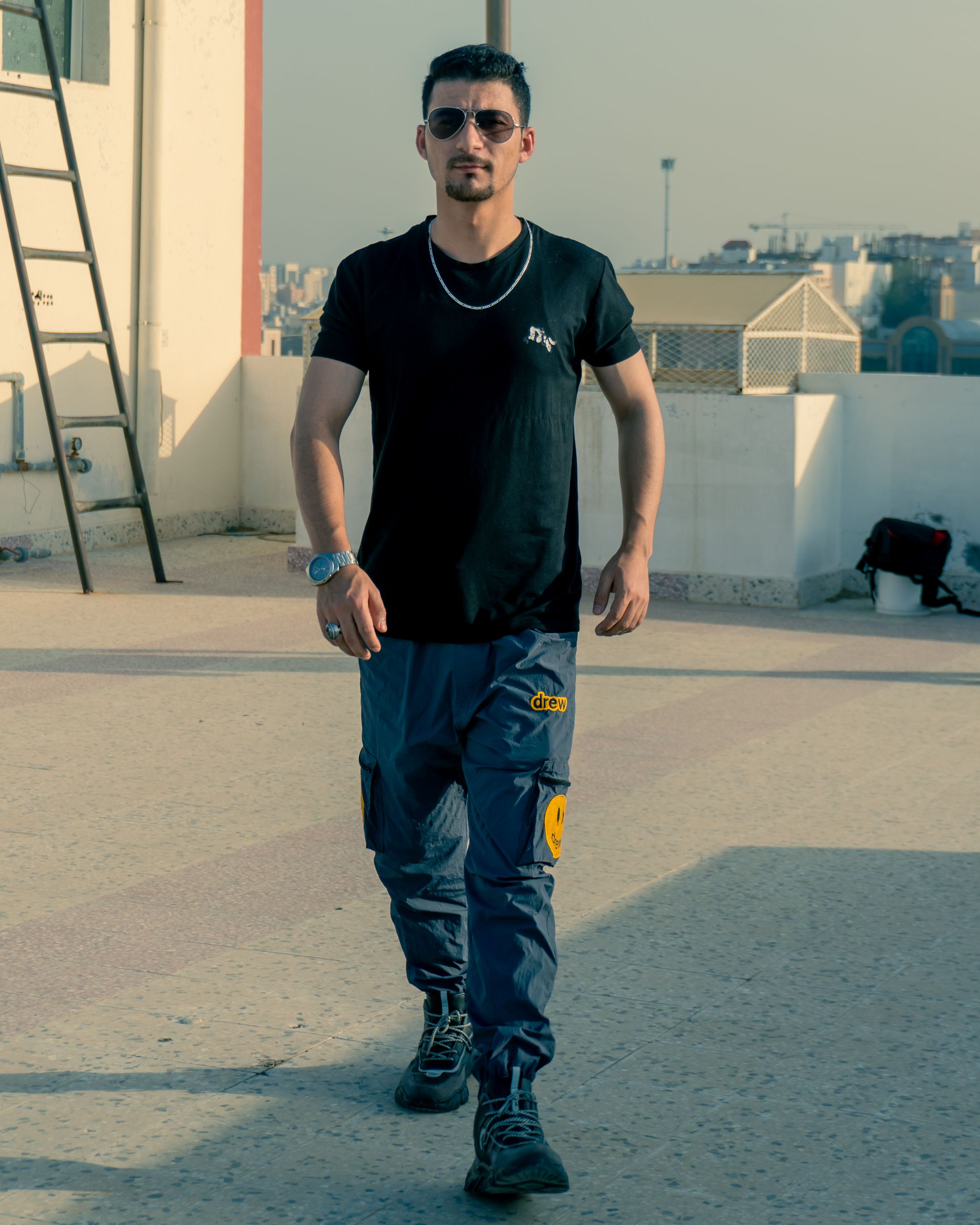 Stylish man posing on a rooftop terrace in summer, wearing sunglasses and casual outfit.