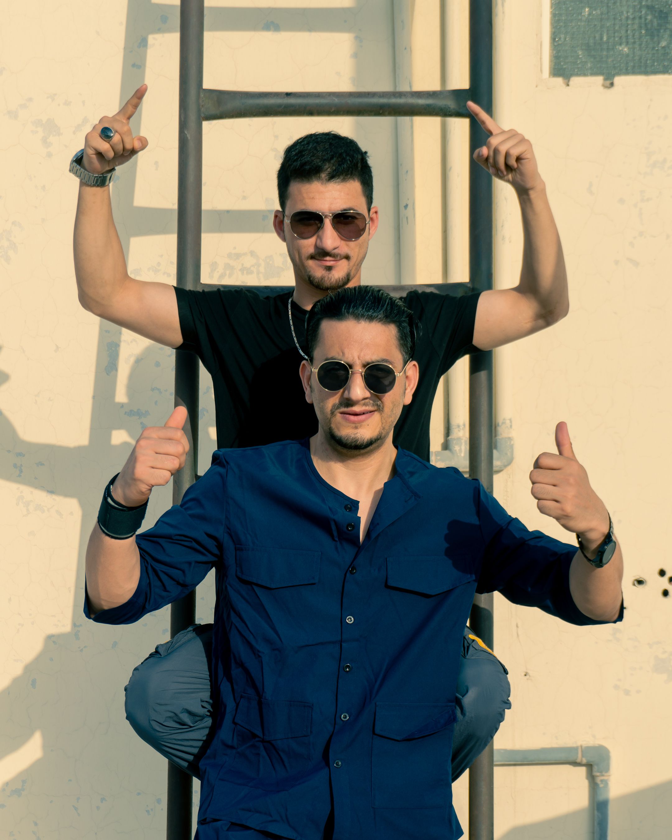 Two men in sunglasses posing confidently on a ladder outdoors in summer sunlight.
